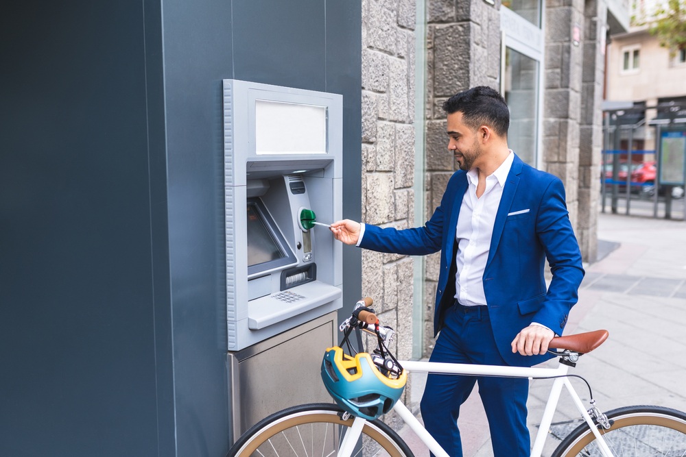 A man with a bike inserts a card into an ATM while smiling. 