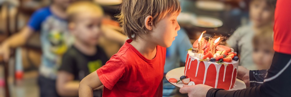 A boy blows out candles on a birthday cake held by a children's party services organizer. 