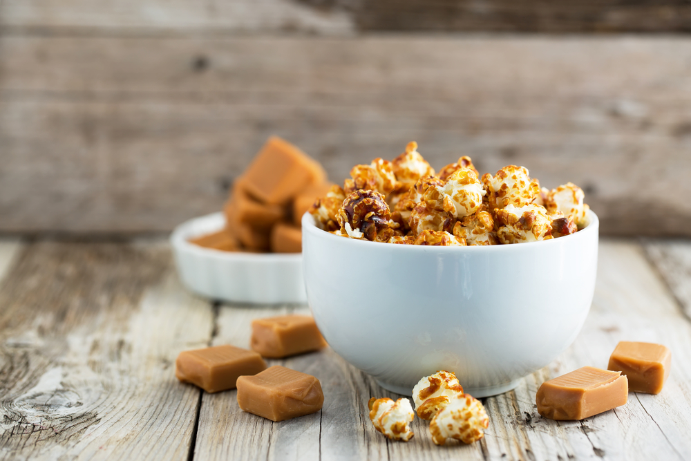 A white bowl of caramel popcorn sits on a table, surrounded by blocks of caramel. 