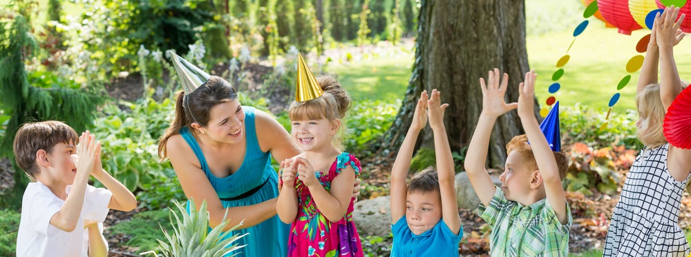 A woman in a birthday hat leads a group of children in a party activity. 