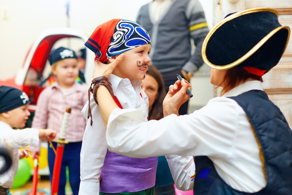 A woman in a pirate outfit paints a mustache on a child attending a party.