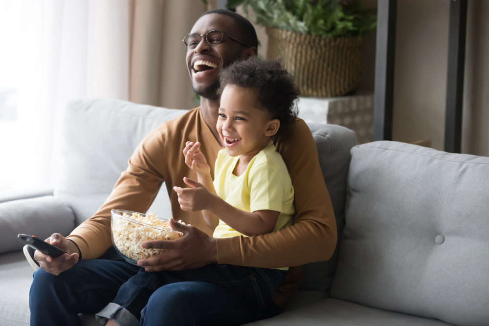 A man holding a remote control sits on a couch with a child on his lap, eating popcorn.