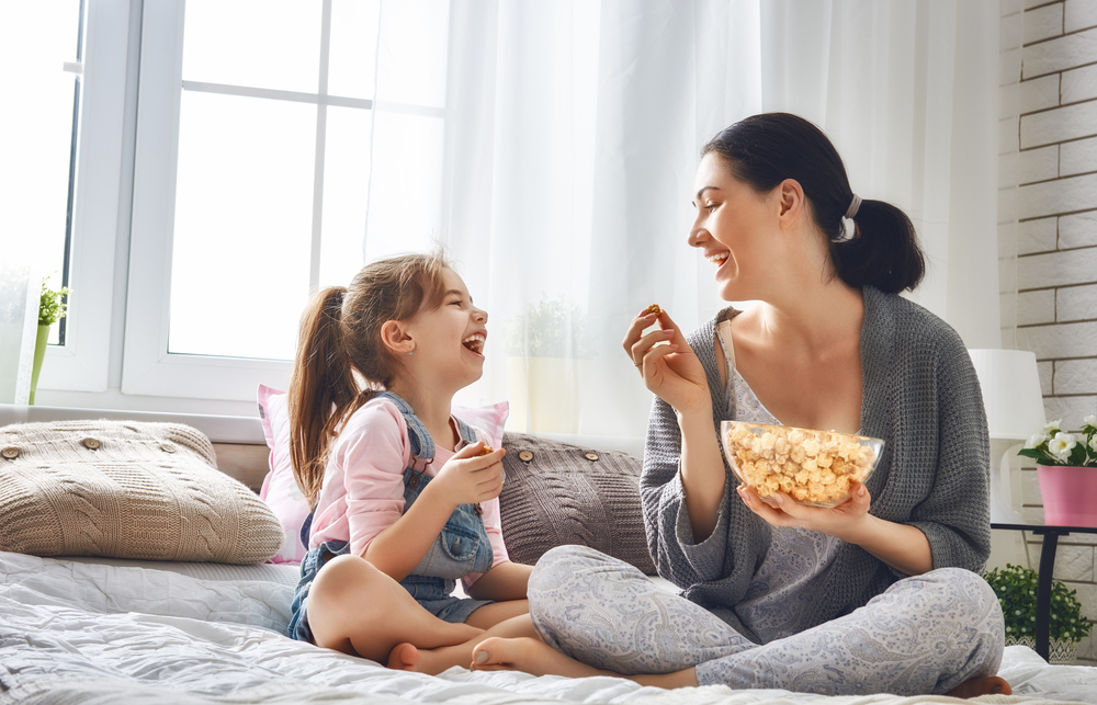 A woman and a child sit on a bed eating popcorn from a bowl, smiling and happy.