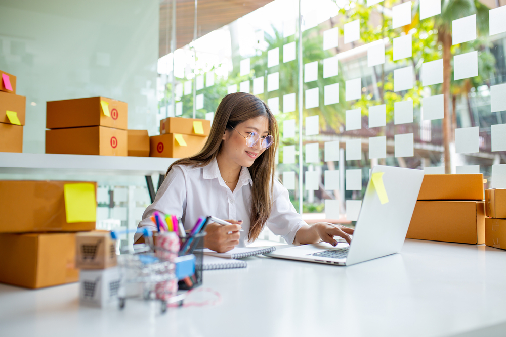 A woman sits in front of a laptop taking notes with a pen and paper and surrounded by boxes of products.