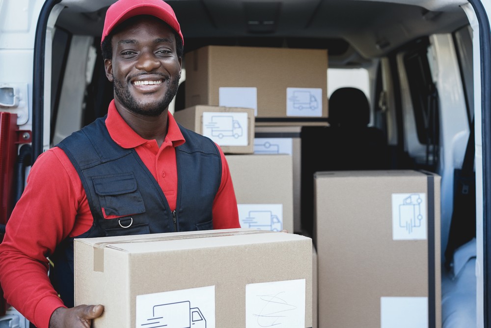 A smiling man holds a box behind a box truck.