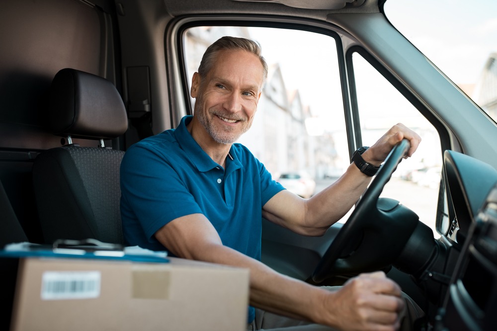 A man smiling with his hand on the gear shift sits in the driver's seat of a box truck.