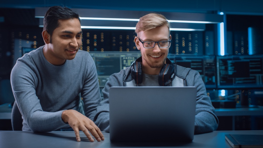 Two men lean over a laptop and laugh.