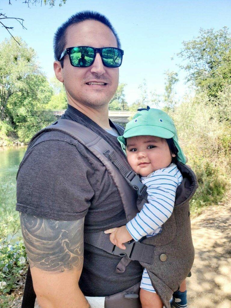 man with a baby strapped to his chest going for a hike in sun glasses and a hat