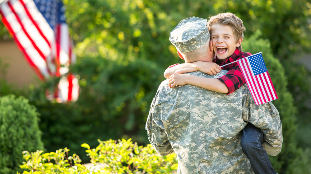 A man in a military uniform holds his smiling son, who is waving a small American flag.