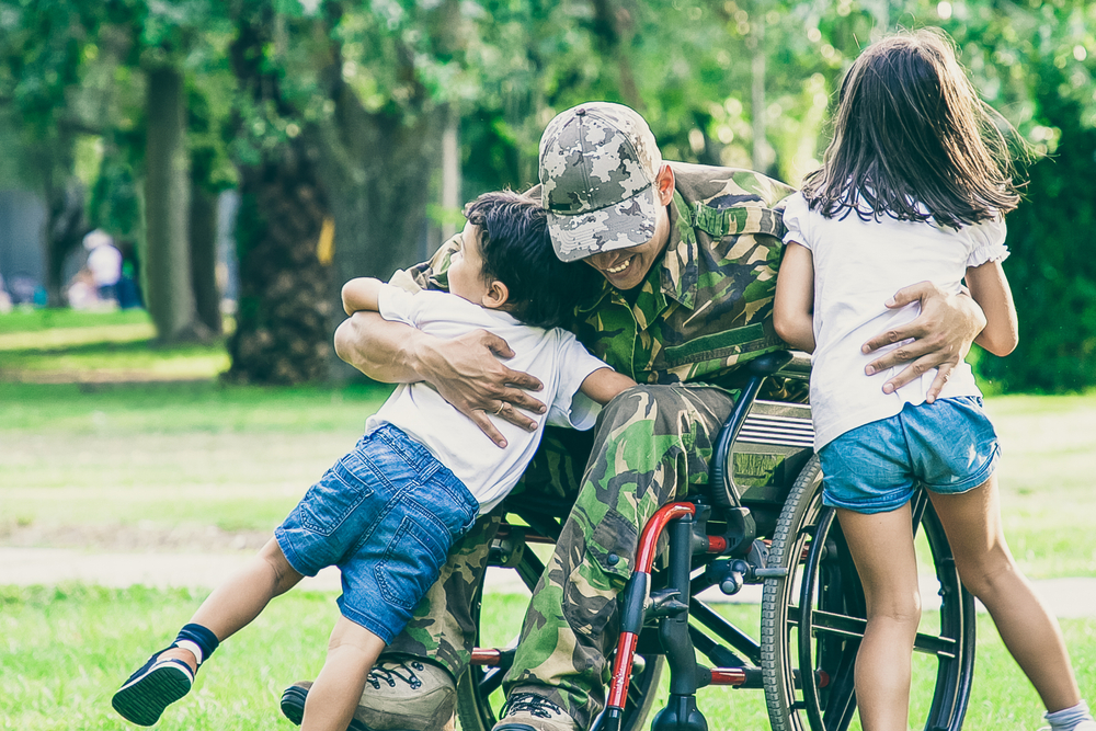 A man in a wheelchair wearing military-style clothing hugs his children.