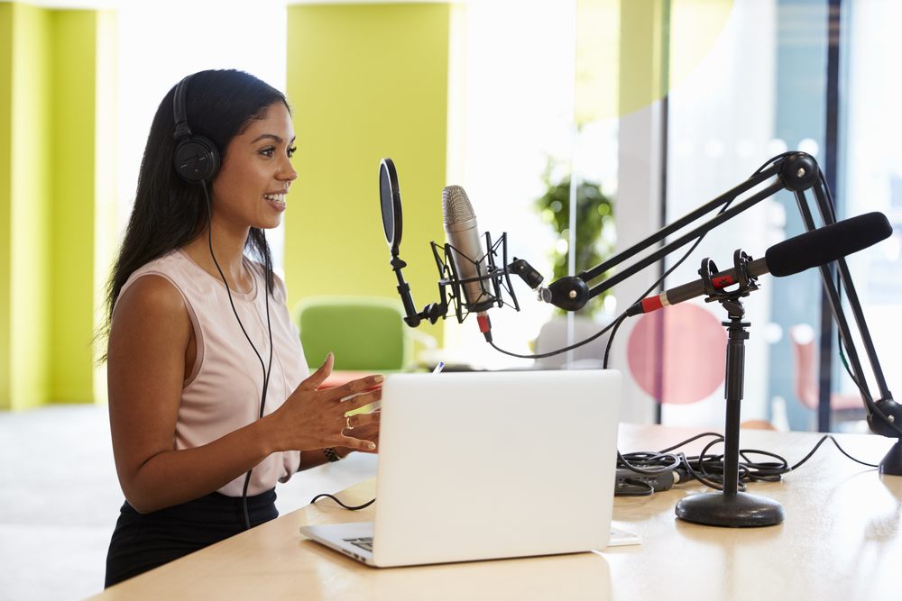 A woman near a laptop and a microphone records a podcast.