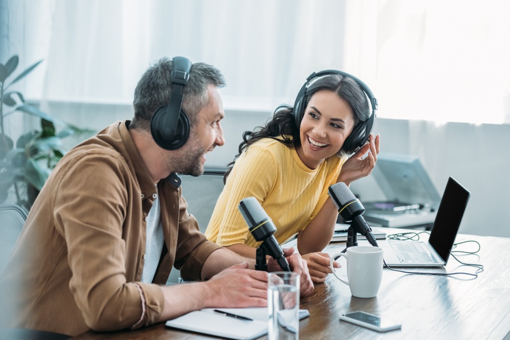 A man and a woman laugh as they record a podcast in front of microphones.