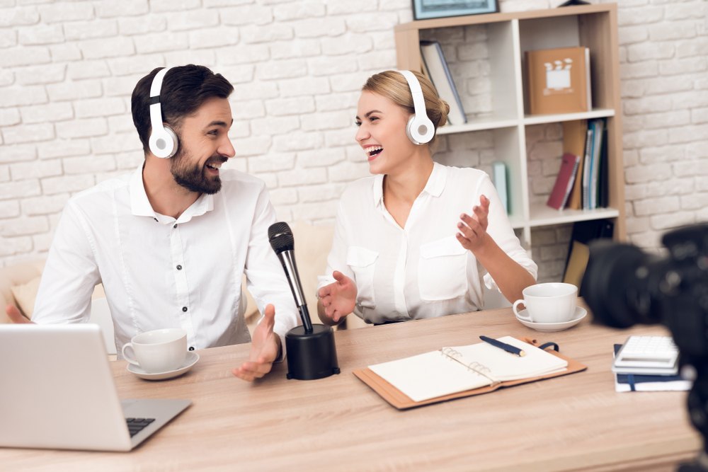 A man and a woman drink coffee and laugh next to a podcast microphone.