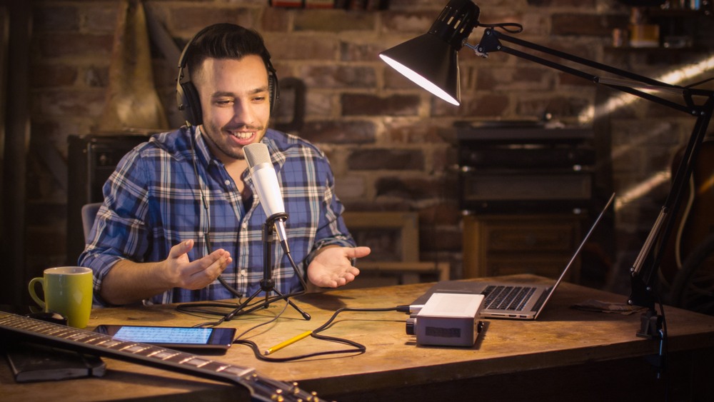 A man in a plaid shirt sits in front of a microphone with a guitar on the table. 