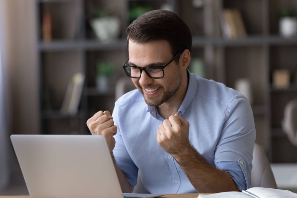 Just won a bid on a freelancer site. young man with glasses sitting in front of a laptop computer looking happy.