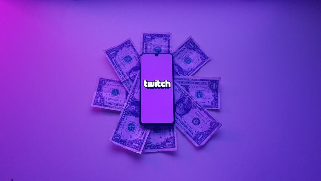 twitch logo on mobile phone resting on pile of dollar bills with purple background and overlay